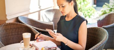 Genç iş kadını, akıllı telefondan not defterine kahve molasında çalışırken ona bakıyor.