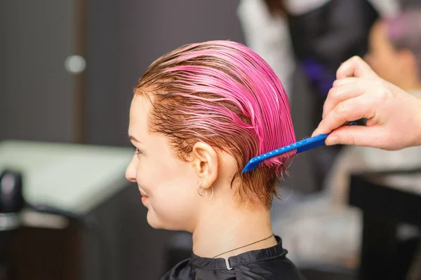 Young woman receiving hair treatment after pink coloring by hand of male hairdresser in hair salon