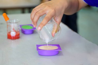 A student pours a creamy liquid from a beaker into a purple mold, for making soap. Other colorful beakers and molds are visible, indicating a hands-on chemistry experiment in a classroom.                               