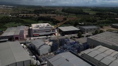 İnsansız hava aracı görüntüleri sanayi bölgesini tarıyor. Depolar, metal raf depolama sistemleri ve ortada bir inşaat vinci var.