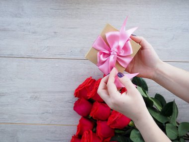 Güllerle tahta bir masada hediye hazırlıyorum. Kırmızı güllerin arka planında kurdele ile sarılmış bir hediye tutan kadın el, metnin altındaki boşluk.