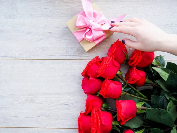 Preparing a gift, on a wooden table, with roses. Female hand holding a gift, wrapped with ribbon, on a background of red roses, the space under the text.