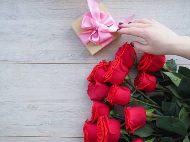Güllerle tahta bir masada hediye hazırlıyorum. Kırmızı güllerin arka planında kurdele ile sarılmış bir hediye tutan kadın el, metnin altındaki boşluk.