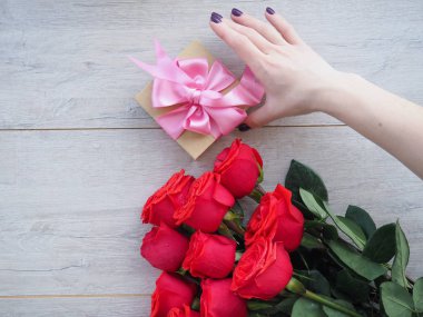 Güllerle tahta bir masada hediye hazırlıyorum. Kırmızı güllerin arka planında kurdele ile sarılmış bir hediye tutan kadın el, metnin altındaki boşluk.