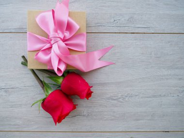 Güllerle tahta bir masada hediye hazırlıyorum. Kırmızı güllerin arka planında kurdele ile sarılmış bir hediye tutan kadın el, metnin altındaki boşluk.