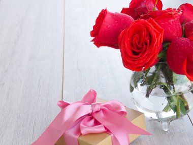 Güllerle tahta bir masada hediye hazırlıyorum. Kırmızı güllerin arka planında kurdele ile sarılmış bir hediye tutan kadın el, metnin altındaki boşluk.