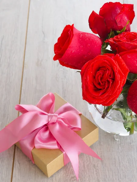 De voorbereiding van een geschenk, op een houten tafel, met rozen. Vrouwelijke hand houden een geschenk, verpakt met lint, op een achtergrond van rode rozen, de ruimte onder de tekst. — Stockfoto