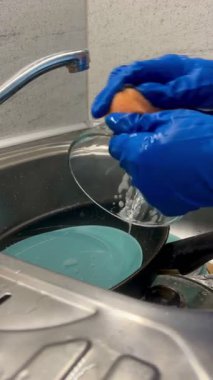 Mutfak lavabosunda bulaşık yıkayan kişi sünger ve eldivenle günlük ev işlerini kapatıyor..