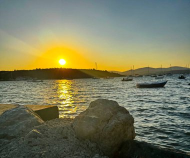 Ufkun üzerindeki günbatımının merkezsel görüntüsü, deniz dalgaları ve kayalık kıyılara altın ışık saçıyor. Arka planda yat ve marinayla sakin bir yaz akşamı..