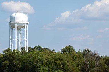 Arka planda mavi gökyüzü olan beyaz bir su kulesi. Beyaz su kulesi dağınık bulutlu mavi bir gökyüzüne karşı duruyor. Kulede silindirik destekli bacakları olan küresel bir tavan var. Aşağıda, ağaçlar gökyüzü ile zıtlık yaratır.. 