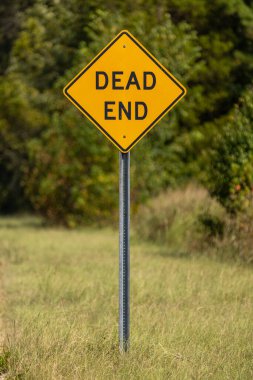 Arka planda yeşil ağaçlar olan çıkmaz bir yol tabelası. Ön planda metal bir direğe monte edilmiş sarı elmas şekilli 