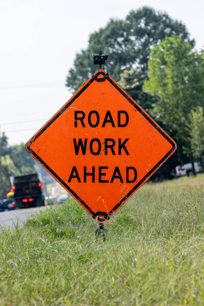 Оранжевый бриллиант в форме предупреждающего знака указывает на дорожные работы впереди. Оранжевая бриллиантовая вывеска гласит "Дорога вперед" жирными черными буквами. Вдоль дороги с видимым движением и в окружении травы. 
