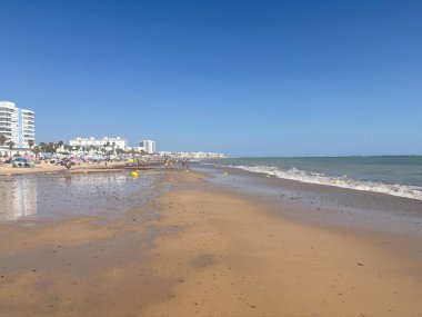 Spain: Golden sands of Playa de la Costilla in Rota, with calm Atlantic waters, promenades, sunbathers, and vibrant chiringuitos under clear sunny skies.