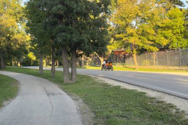 Yeşil ağaçlar ve akşam güneşiyle çevrili park yolunda dört tekerli bisiklet süren iki kişi, dışarıda dinlenme ve boş vakitlerin keyfini çıkarıyorlar..