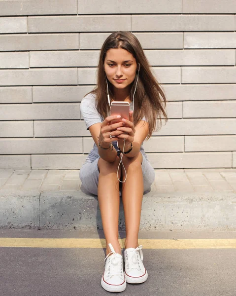 Cheerfully beautiful young woman in fashionable clothes, while enjoying the online playlist on headphones and reading sms with good news on the smartphone.