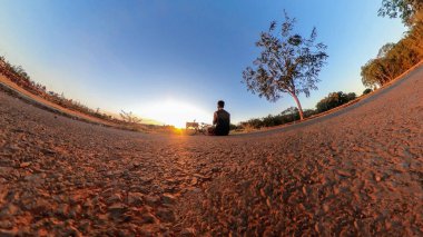 Parkta ve gün batımında maske takan bir adamın açık hava draması. Ağaçlı, yumuşak mavi gökyüzü ve günbatımı olan parkta bisikletli bir adam.. 