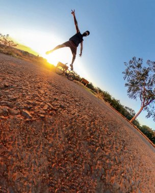 Parkta ve gün batımında maske takan bir adamın açık hava draması. Ağaçlı, yumuşak mavi gökyüzü ve günbatımı olan parkta bisikletli bir adam.. 