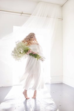 Kız beyaz ışık elbise ve kıvırcık saçlı, pencere, saflık ve masumiyet yakınındaki evde çiçekli kadın portresi. Kıvırcık sarışın romantik bak, güzel gözler. Elinde beyaz kır çiçekleri