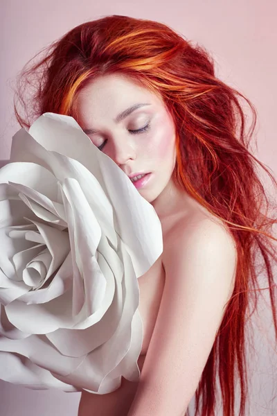 Nude woman with red hair hugs a big paper flower. Beautiful long hair girl, beautiful body. Artificial paper rose flower in the hands of a woman. Hair and body care, perfect skin