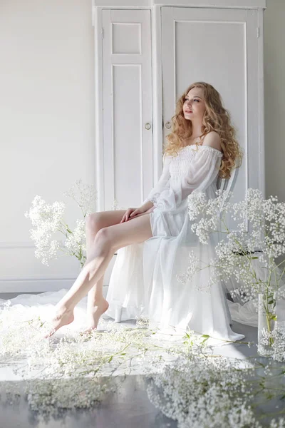 Curly blonde romantic look, beautiful eyes. White wildflowers in hands. Girl white light dress and curly hair, portrait of woman with flowers at home near the window, purity and innocence