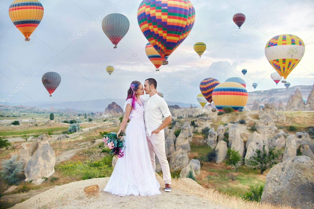 Couple in love stands on background of balloons in Cappadocia. Man and a woman on hill look at a large number of flying balloons. Turkey Cappadocia fairytale scenery of mountains. Wedding on nature