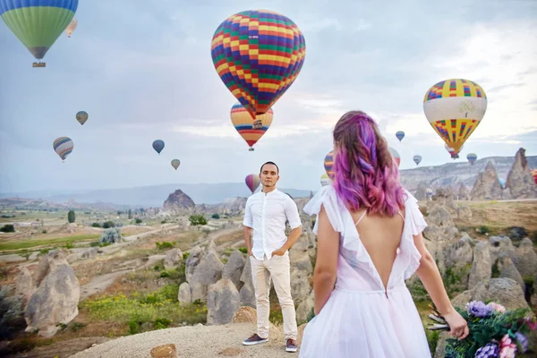 Couple in love stands on background of balloons in Cappadocia. Man and a woman on hill look at a large number of flying balloons. Turkey Cappadocia fairytale scenery of mountains. Wedding on nature