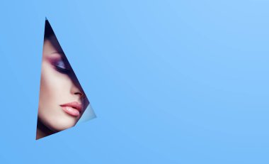 Kadın mavi kağıt, parlak makyaj ve pembe ruj üçgen bir delikten güzel bir görünüme sahip. Reklam kozmetik, profesyonel sanat makyaj, dudak parlatıcısı, parlak renk, mavi arka plan