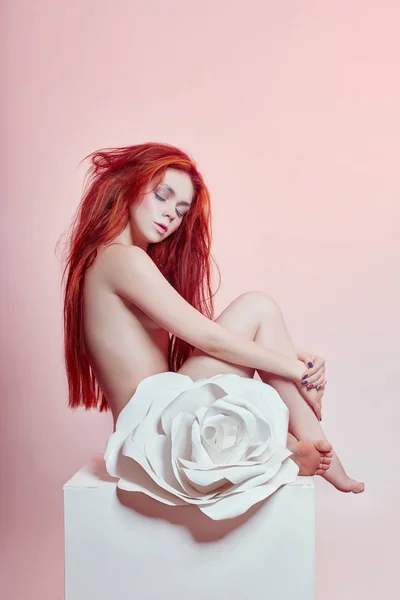 Naked woman with red hair sitting near a large paper flower. Beautiful long hair girl, beautiful body. Artificial paper rose flower on a woman\'s body. Hair and body care, perfect skin