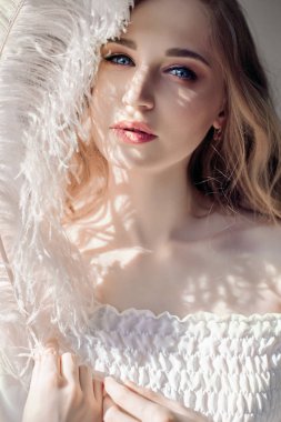 Beyaz ışık elbise ve onun yüz, tüy, saflık ve masumiyet ile kadın portre yakınındaki büyük tüy kıvırcık saçlı kız. Kıvırcık sarışın romantik, güzel gözleri görünüyor. Beyaz devekuşu tüyü elinde