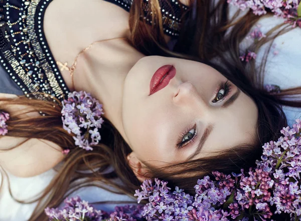 夏の日に公園で紫の花ライラックの枝に横たわってポーズ ドレスで美しい少女 春の太陽の下で自然の中で夢を見る少女の肖像画 顔に複雑な心境 — ストック写真