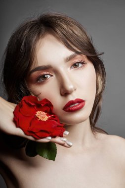 Saçında büyük kırmızı bir çiçek olan kadın. Kahverengi saçlı kız poz gri bir arka plan üzerinde kırmızı çiçekli. Güzel gözleri iri ve doğal makyaj. Uzun kıvırcık saç, mükemmel yüz