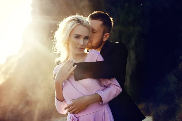 在日落的夏天 男人和一个女人穿着漂亮的衣服拥抱在一起 恋爱中的一对夫妇站在岸边的阳光下的岩石上 在水花中拥抱和亲吻 粉红色连衣裙 — 图库照片