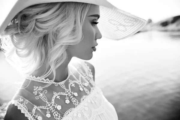 Woman in a big white hat stands near the sea, a blonde in a hat looking at the lake, summer vacation travel and vacation. Sun rays at sunset