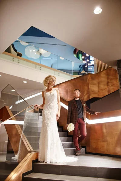 Bride in a chic long dress with a train and the groom stand on big stairs, couple in love hugs on stairs kiss and look at each other. Day of the wedding, couple on the day of registration of marriage