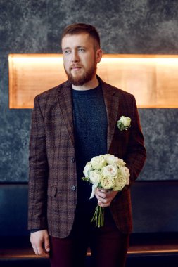 Morning bridegroom. A hipster man in a jacket holding a bouquet of flowers for his beloved. Beautiful bearded man preparing for the wedding ceremony
