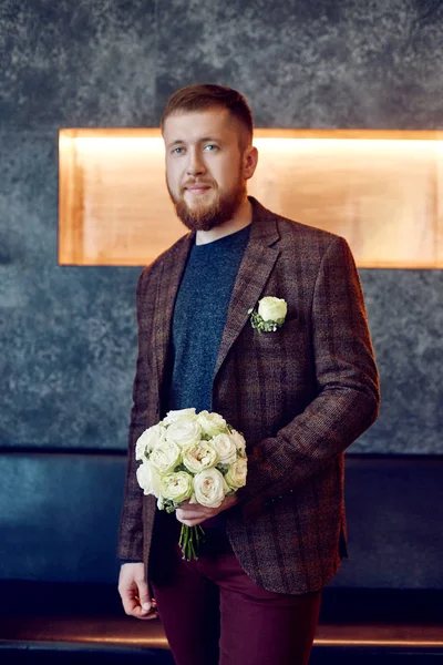 Morning bridegroom. A hipster man in a jacket holding a bouquet of flowers for his beloved. Beautiful bearded man preparing for the wedding ceremony