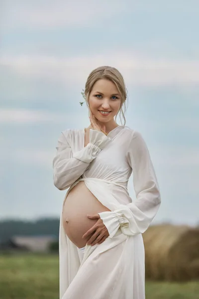 Беременная девушка ходит по полю возле стога сена в длинном белом Докторе — стоковое фото