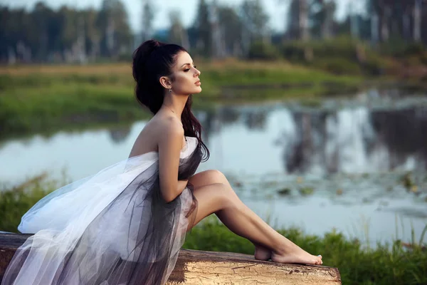 Ragazza in un abito lungo vicino al lago si siede su una vecchia barca, una bella — Foto Stock