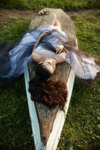 Menina em um vestido longo perto do lago senta-se em um barco velho, um beauti — Fotografia de Stock