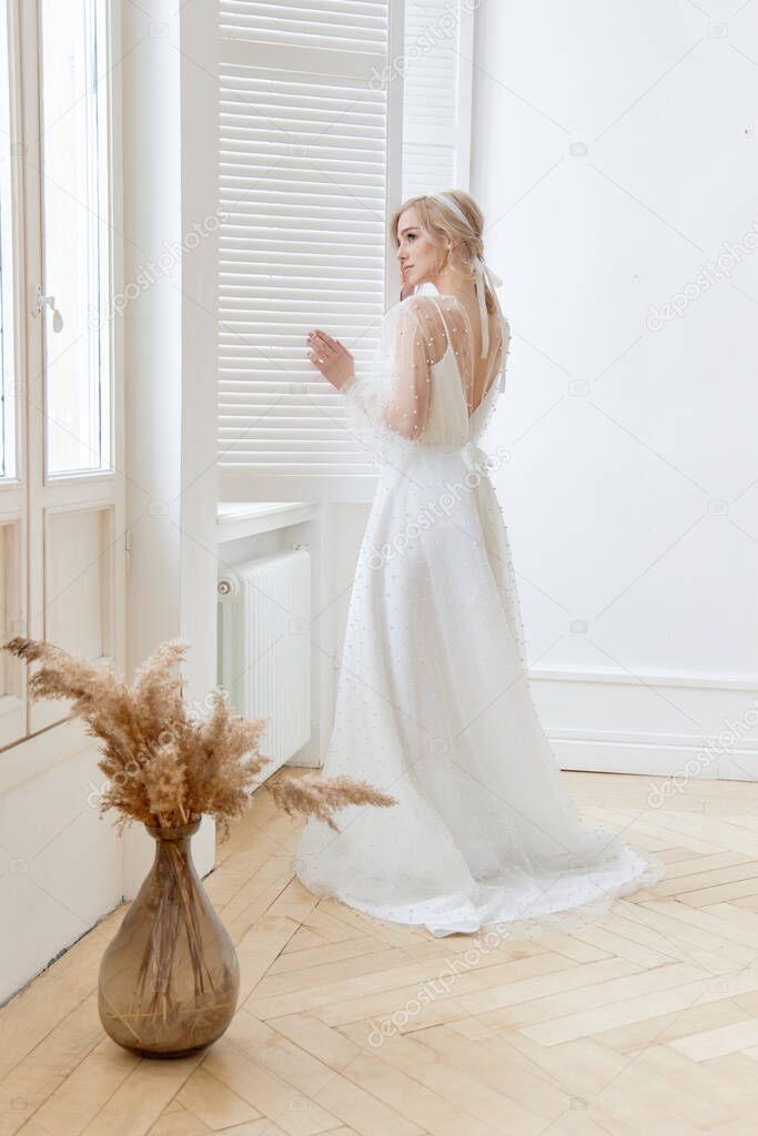 Romantic portrait of a woman near the window in a beautiful long white dress at home. The girl is blonde with blue eyes and beautiful makeup on her face. Natural cosmetics