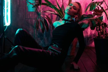 Moda kadını tropikal neon ışıkta yerde oturuyor. Islak saç, mükemmel vücut ve makyaj. Pembe ve yeşil neon rengi. Güzel kız ve canavar yaprakları