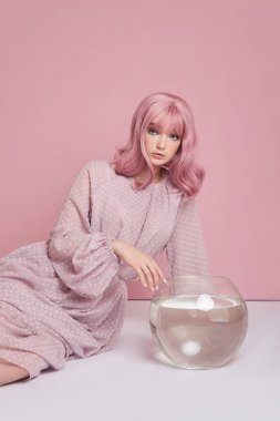 Uzun elbiseli, boyalı pembe saçlı kız yuvarlak bir akvaryumla yerde oturuyor. Pembe duvarda saç boyalı bir portre kadın. Mükemmel saç stili ve saç şekli