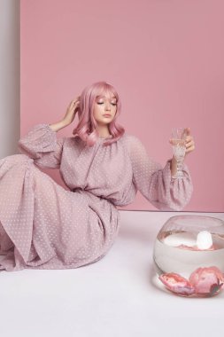 Uzun elbiseli, boyalı pembe saçlı kız yuvarlak bir akvaryumla yerde oturuyor. Pembe duvarda saç boyalı bir portre kadın. Mükemmel saç stili ve saç şekli