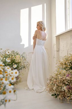 Uzun beyaz gelinlikli romantik seksi kadın papatya çiçekleri içinde pencerenin yanında duruyor. Mükemmel vücutlu sarışın kız.