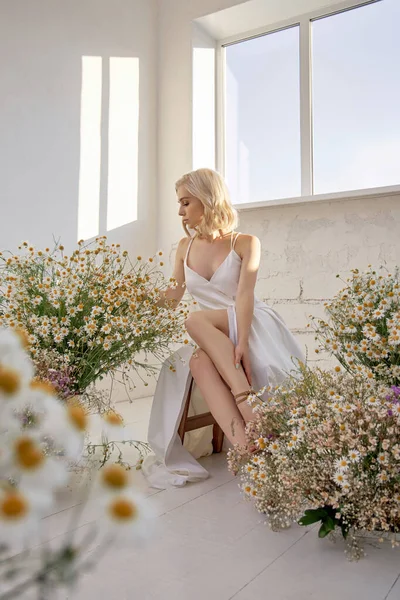 美しい白いドレスのセクシーなブロンドの女性が野生の花の花束の前のウィンドウの近くに座っている ロマンチックな女の子とともに美しい自然の化粧 — ストック写真