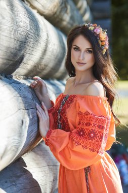 Beautiful Slavic woman in an orange ethnic dress and a wreath of flowers on her head. Beautiful natural makeup. Portrait of a Russian girl clipart
