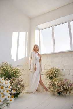 Ceketli ve uzun beyaz elbiseli romantik seksi kadın papatya çiçekleri içinde pencerenin yanında duruyor. Mükemmel vücutlu sarışın kız.