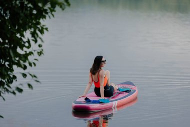Bir kürekçi, sabah güneşi suyun üzerine yumuşak bir ışık saçarken yeşilliklerle çevrili sakin bir gölde dinlenir. Sakinlik ve yalnızlık bu huzurlu deneyimi tanımlar..