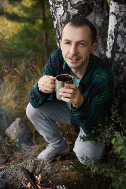 Adam, dingin bir ormanda, bir kamp ateşinin yanında oturmuş elinde sıcak bir içecekle dolu metal bir bardak tutarken çevresi yeşillik ve doğa sesleri ile çevrili..