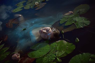 Zarif hatlara sahip bir kadın yemyeşil nilüferlerle çevrili bir gölette huzur içinde yüzüyor. Yumuşak öğleden sonra ışığı sahnenin sakin atmosferini arttırır..
